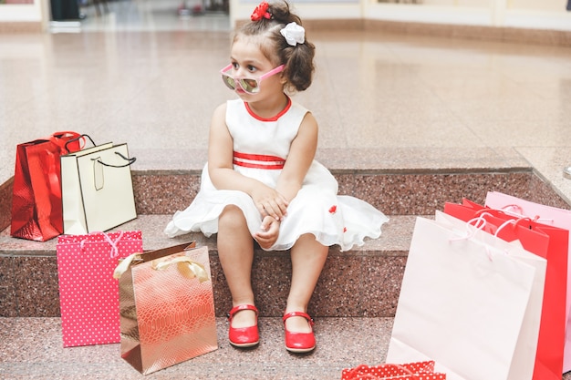 Mała dziewczynka w okularach przeciwsłonecznych siedzi na schodach w centrum handlowym z zakupami