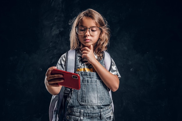 Mała Dziewczynka W Okularach Przewija Telefon Komórkowy W Ciemnym Studiu Fotograficznym.