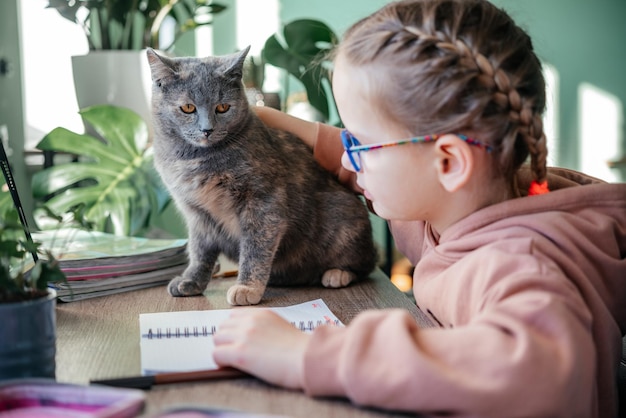 Mała dziewczynka w okularach ze swoim kotem ma lekcję online podczas edukacji na odległość krępowania...