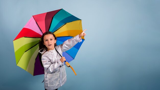 Mała dziewczynka w płaszczu przeciwdeszczowym trzyma wielokolorowy parasol i wskazuje ręką na niebieskim tle z kopią przestrzeni