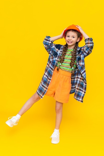 Mała dziewczynka w pomarańczowym kasku budowlanym trzyma głowę i uśmiecha się szeroko radośnie Dziecko przygotowuje się do naprawy w pokoju dziecinnym W kęsie Pionowe żółte odosobnione tło