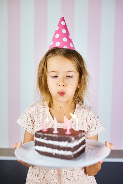 Mała dziewczynka w różowej nakrętce zdmuchuje świeczki na urodzinowym torcie czekoladowym na jej przyjęciu urodzinowym w domu. Urodziny dziecka portretowego.