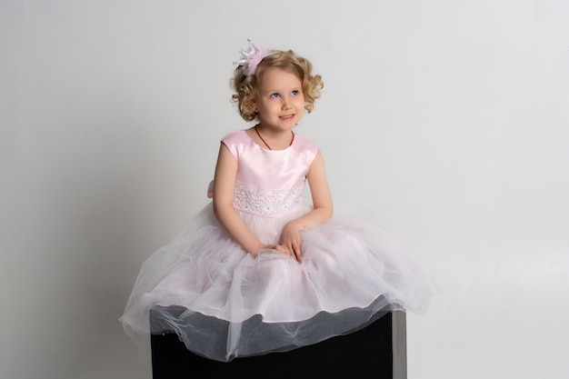 Mała dziewczynka w różowej sukience i koronie siedzi na czarnym sześcianie na białym tle.