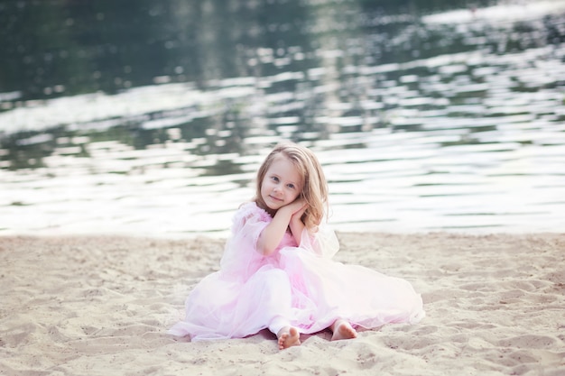 Mała dziewczynka w różowej sukience siedzi na piasku, ciesząc się wakacjami i rodzinną wycieczką. Spoczywaj na morzu. Zabawne dzieci. Urocza śliczna młoda dziewczyna bawić się na plaży przy zmierzchem