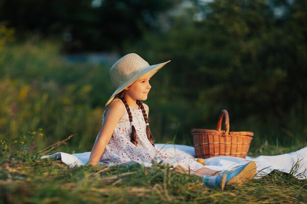 mała dziewczynka w słomkowym kapeluszu i sukience siedząca na zielonej trawie