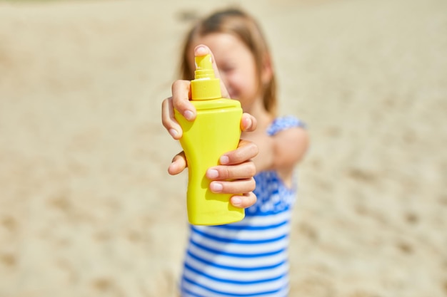 Mała dziewczynka w stroju kąpielowym trzymać w ręku żółtą butelkę ochrony przeciwsłonecznej stojąc na plaży, ochrona skóry krem do opalania. Letnie wakacje.