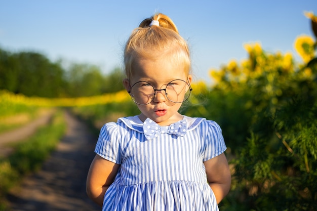 Mała dziewczynka w sukience i okularach na tle słonecznika. Emocjonalna twarz.