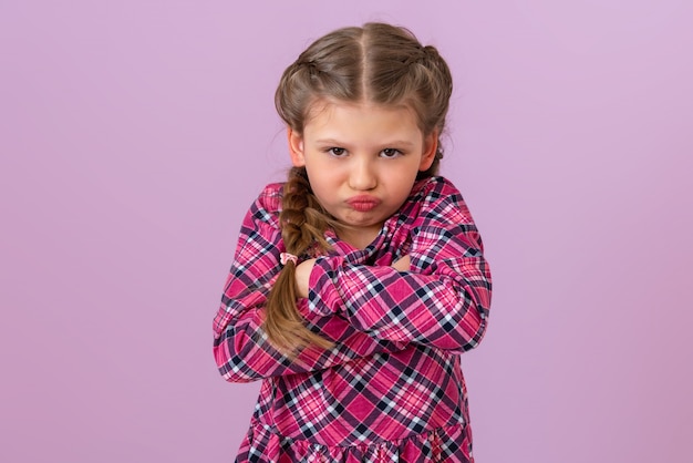 Mała dziewczynka w sukience w kratę iz warkoczykami na głowie trzyma ręce przy piersiach i wygląda na bardzo urażoną.