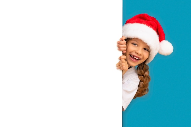 Mała dziewczynka w świątecznej czapce wygląda zza reklamy.