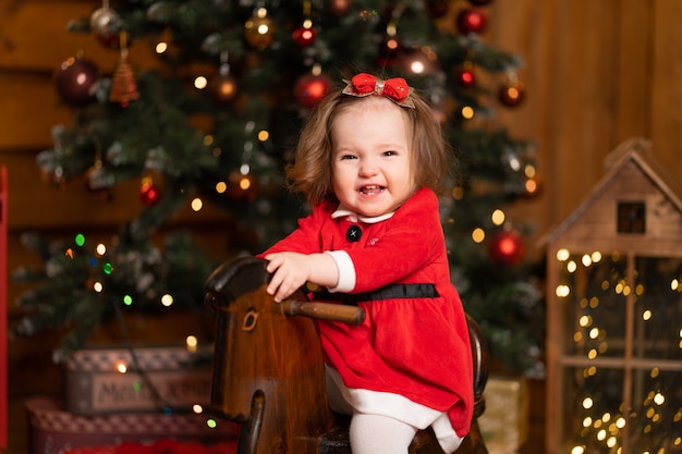 Mała dziewczynka w świątecznej czerwonej sukience na koniku na biegunach drewnianej huśtawki