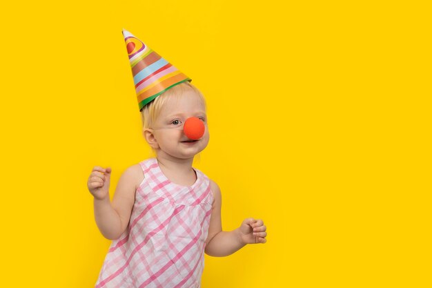 Mała dziewczynka w świątecznym kapeluszu i nosie klauna na żółtym tle.