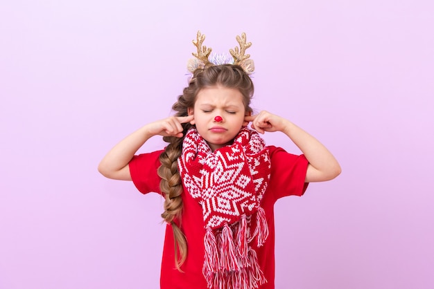 Mała dziewczynka w świątecznym stroju renifera iz ciepłym szalikiem włożyła palce do uszu i zamknęła oczy.