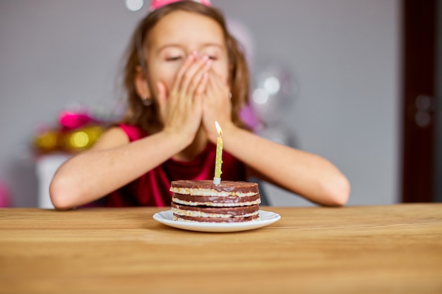 Zdjęcie mała dziewczynka w urodzinowej czapce wypowiada życzenie, patrząc na tort urodzinowy
