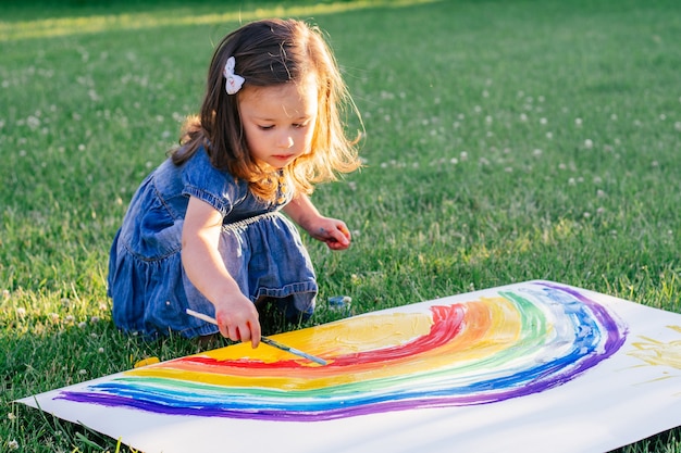 Mała dziewczynka w wieku 2-4 lat maluje tęczę i słońce na dużej kartce papieru, siedząc na zielonym trawniku