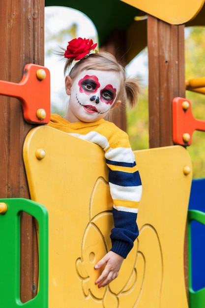 Zdjęcie mała dziewczynka w wieku przedszkolnym z pomalowaną twarzą wspięła się na zjeżdżalnię na placu zabaw, świętuje halloween lub meksykański dzień zmarłych.