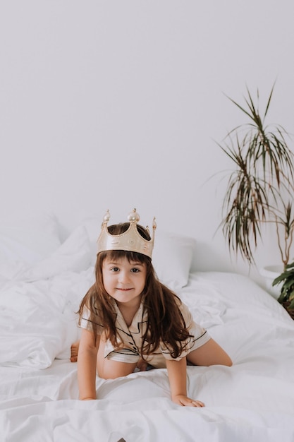 mała dziewczynka w złotej piżamie i koronie w łóżku w domu z białą pościelą. poranek małego