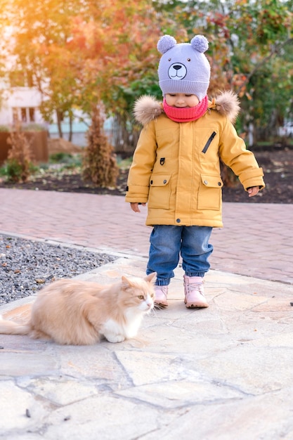Mała dziewczynka w żółtej kurtce bawi się z czerwonym kotem na jesiennym spacerze