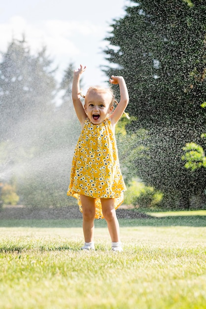 Zdjęcie mała dziewczynka w żółtej sukience z podniesionymi rękami stoi przed zraszaczem, który jest spryskiwany wodą.