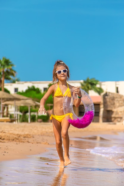 Mała dziewczynka w żółtym bikini i różowym nadmuchiwanym pierścieniem biegnie po plaży