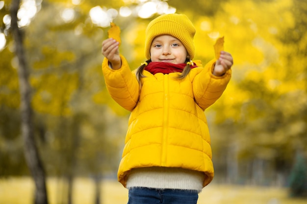 Mała dziewczynka w żółtym płaszczu i czapce, uśmiecha się do kamery w pięknym jesiennym parku, trzymając w dłoniach liście. Sezon jesienny, portret dziecka, koncepcja szczęśliwego dzieciństwa.