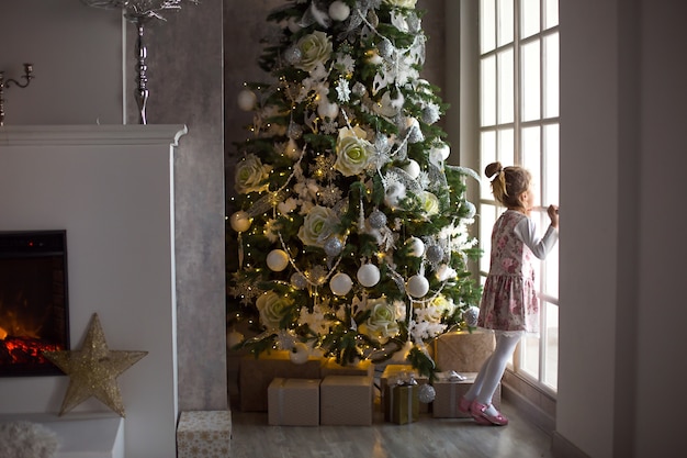 Mała dziewczynka wygląda przez duże okno w pobliżu choinki. Czekam na cudowny, świąteczny wystrój w kolorze białym w salonie domu. Nowy rok, bajka i magia, dziecięce marzenia