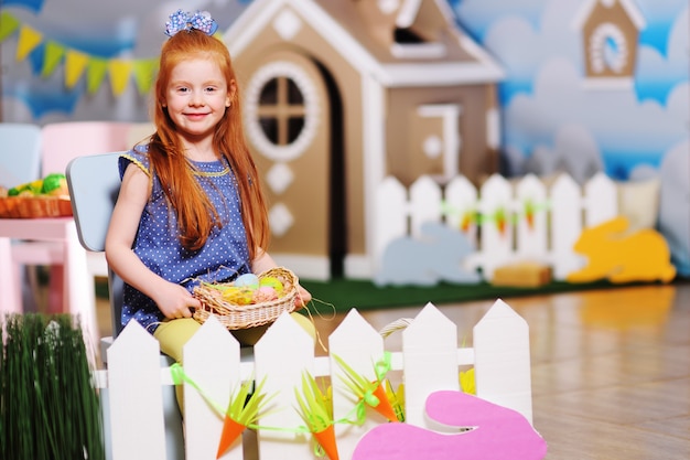 Mała dziewczynka z czerwonymi włosami z wielkanocnym koszem na białym ogrodzeniu i lalki zabawki domem. Przygotowanie do obchodów Wielkanocy