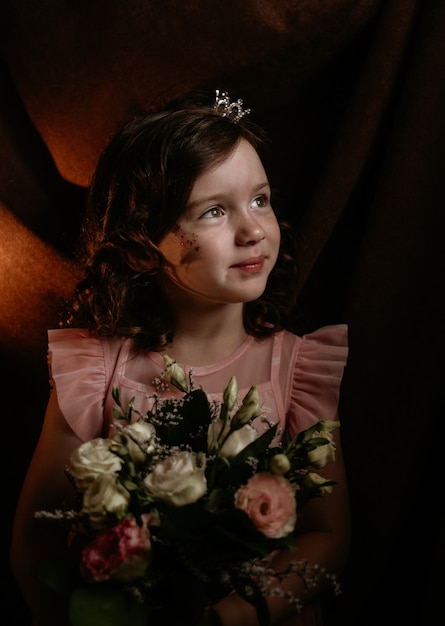 Mała dziewczynka z kręconymi włosami trzymająca bukiet kwiatów róży odwraca wzrok na brązowym tle