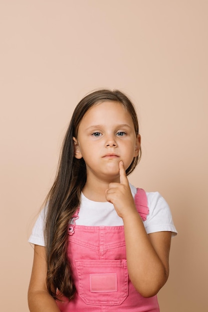 Mała dziewczynka z lśniącymi jasnymi oczami patrząca na palec wskazujący aparat na obszarze podbródka twarzy z zamyślonym spojrzeniem, ubrana w jasny różowy kombinezon i białą koszulkę na beżowym tle