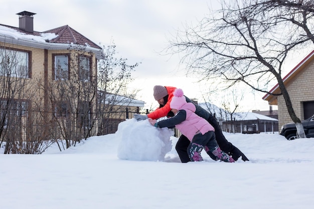Mała dziewczynka z matką robi masywną śnieżną kulę do budowania bałwana na podwórku wieczorem z drzewami i domem w tle Rodzice spędzają czas z dziećmi