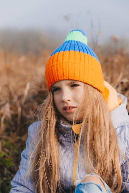 Mała dziewczynka z pięknymi blond włosami siedzi na polu w pobliżu rolki siana. portret dziecka w czapce z dzianiny.