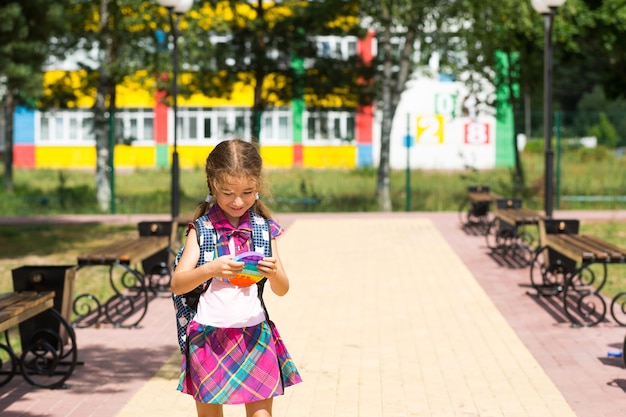 Mała dziewczynka z plecakiem i w szkolnym mundurku na szkolnym boisku bawi się zabawką pop it. Powrót do szkoły, 1 września. Uczeń odpoczywa po lekcjach. Szkolnictwo podstawowe, klasa podstawowa dla ucznia