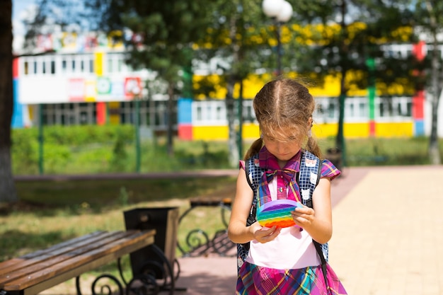 Mała dziewczynka z plecakiem i w szkolnym mundurku na szkolnym boisku bawi się zabawką pop it. Powrót do szkoły, 1 września. Uczeń odpoczywa po lekcjach. Szkolnictwo podstawowe, klasa podstawowa dla ucznia