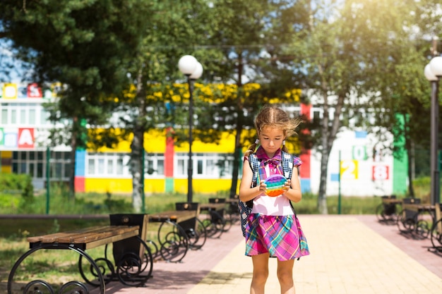 Mała dziewczynka z plecakiem i w szkolnym mundurku na szkolnym podwórku bawi się zabawką pop it.