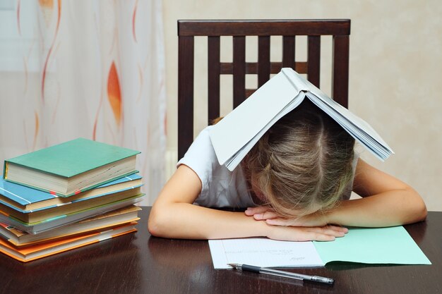 Mała dziewczynka z podręcznikami wygląda na zmęczoną i opiera się na rękach, siedząc przy stole i studiując