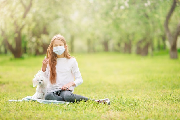 Mała dziewczynka z psem jest ubranym ochronną medyczną maskę dla zapobiega wirusa outdoors w parku