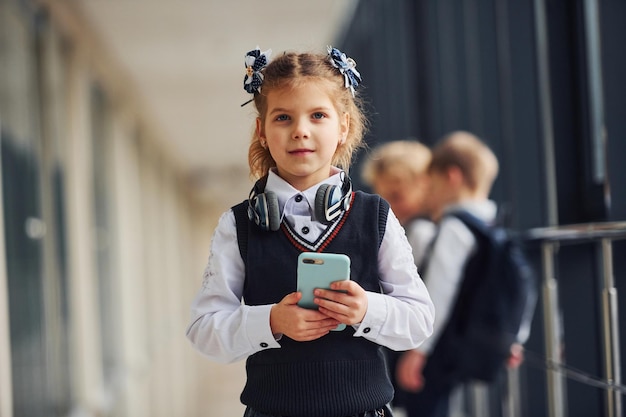 Mała dziewczynka z telefonem stojąca przed dziećmi szkolnymi w mundurach, które razem na korytarzu Koncepcja edukacji