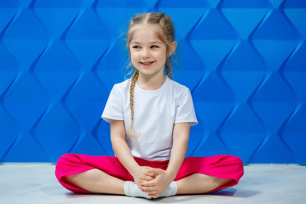 Mała dziewczynka z warkoczykami w białej koszulce na niebieskim tle. Siedzi w pozycji lotosu na podłodze, uśmiecha się i macha rękami.