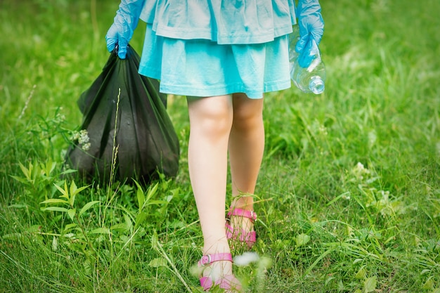 Mała dziewczynka z zmiętą plastikową butelką i workiem na śmieci w rękach podczas sprzątania śmieci w parku