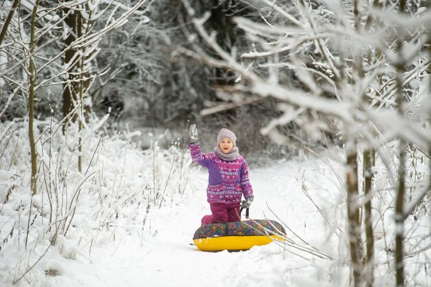 Mała dziewczynka zimą w fioletowych ubraniach i nadmuchiwanym kółku spaceruje po ulicy w pokrytym śniegiem lesie.