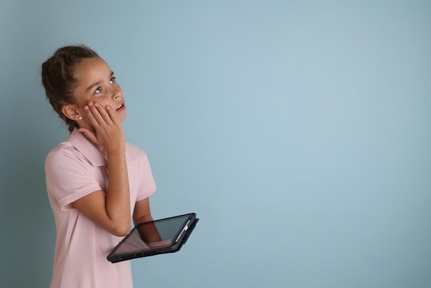 Mała emocjonalna nastolatka w różowej koszuli 11 12 lat na odizolowanym niebieskim tle z tabletem w dłoniach Portret studyjny dzieci 39 lat Umieść tekst, aby skopiować miejsce na napis