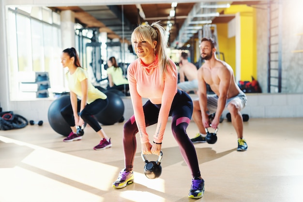 Mała grupa ludzi o zdrowych nawykach wymachuje kettlebell. Wnętrze siłowni, lustro w tle. Mężczyzna