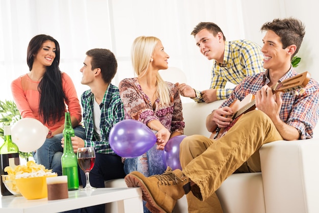 Zdjęcie mała grupa młodych ludzi spędza czas na przyjęciu domowym, rozmawiając ze sobą, podczas gdy ich przyjaciel bawi się grając na gitarze akustycznej.