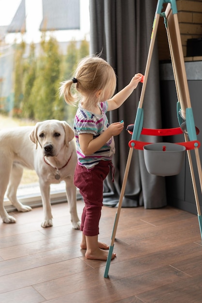 Mała jasnowłosa dziewczynka stoi przy sztalugach przy oknie w pomieszczeniu, a labrador jest w pobliżu