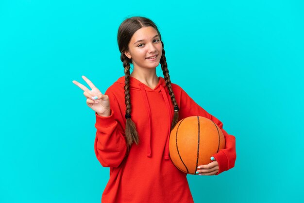 Mała kaukaska dziewczyna gra w koszykówkę na białym tle na niebieskim tle, uśmiechając się i pokazując znak zwycięstwa