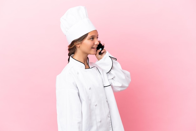 Zdjęcie mała kaukaska dziewczyna kucharza odizolowana na różowym tle, prowadząca rozmowę z telefonem komórkowym