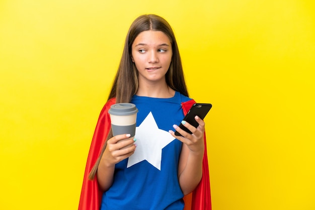 Mała kaukaska dziewczyna superbohatera odizolowana na żółtym tle trzymająca kawę na wynos i telefon komórkowy, myśląc o czymś