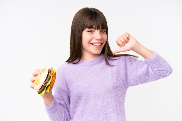 Mała kaukaska dziewczyna trzyma burgera na odosobnionym tle, dumna i zadowolona z siebie