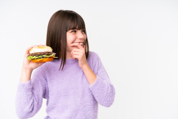 Mała kaukaska dziewczyna trzyma burgera na odosobnionym tle, myśląc o pomyśle i patrząc z boku