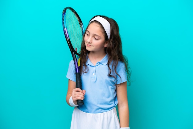 Mała kaukaska dziewczynka gra w tenisa na niebieskim tle patrząc z boku