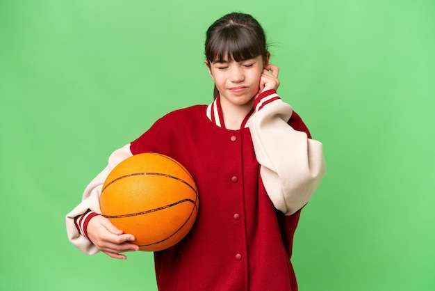 Mała kaukaska dziewczynka grająca w koszykówkę na odosobnionym tle sfrustrowana i zakrywająca uszy